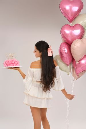 Young woman in white dress holding pink birthday cake and heart-shaped balloons, viewed from behind.