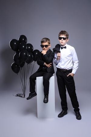 Two children in formal attire and sunglasses posing with black balloons against grey background.