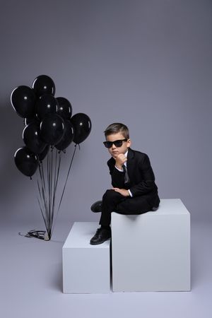 Stylish photo of a child in black suit and sunglasses posing with black balloons on white blocks.