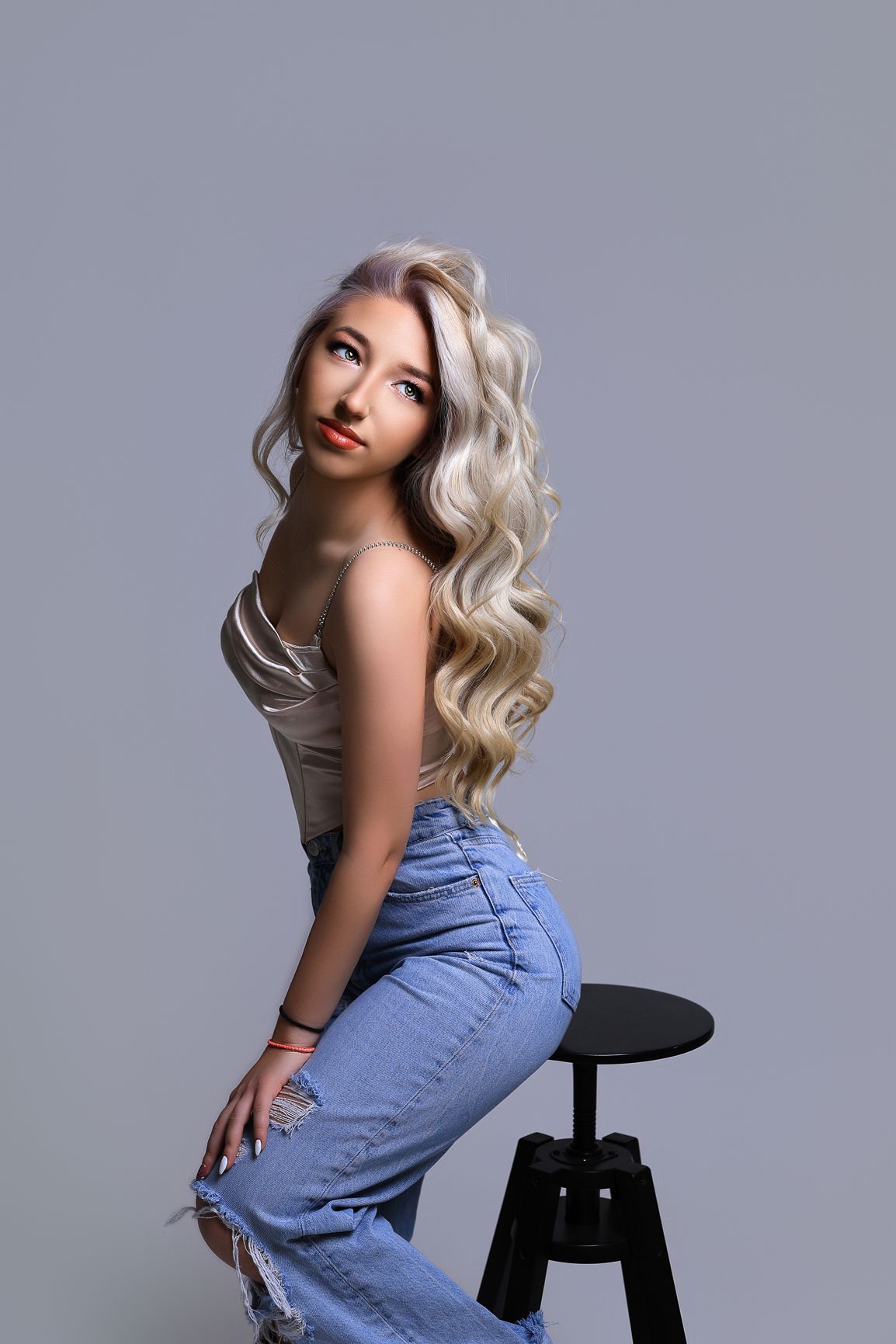 Model in denim poses on black stool for fashion portrait against grey background.