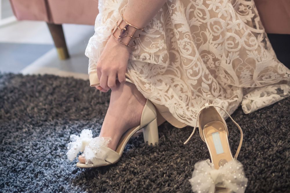 A person in a lace dress puts on elegant white heels with fluffy embellishments while sitting on a dark carpet.