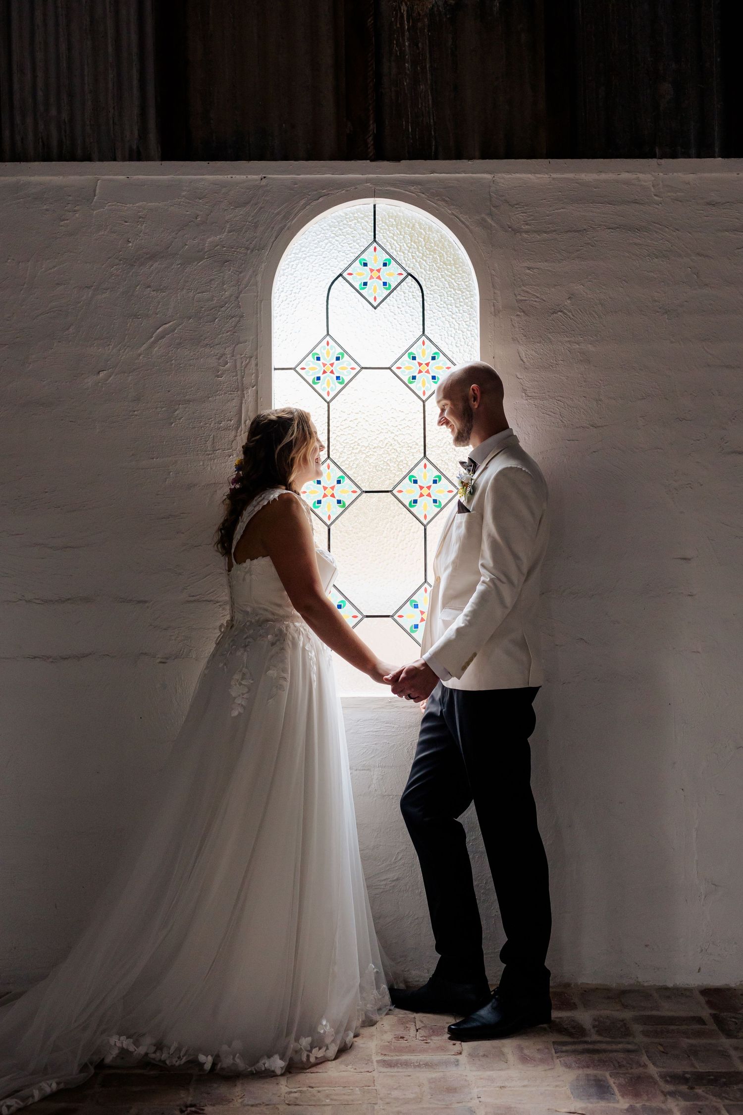 A couple in wedding attire poses together by a bright church window, creating a romantic silhouetted effect against white walls.