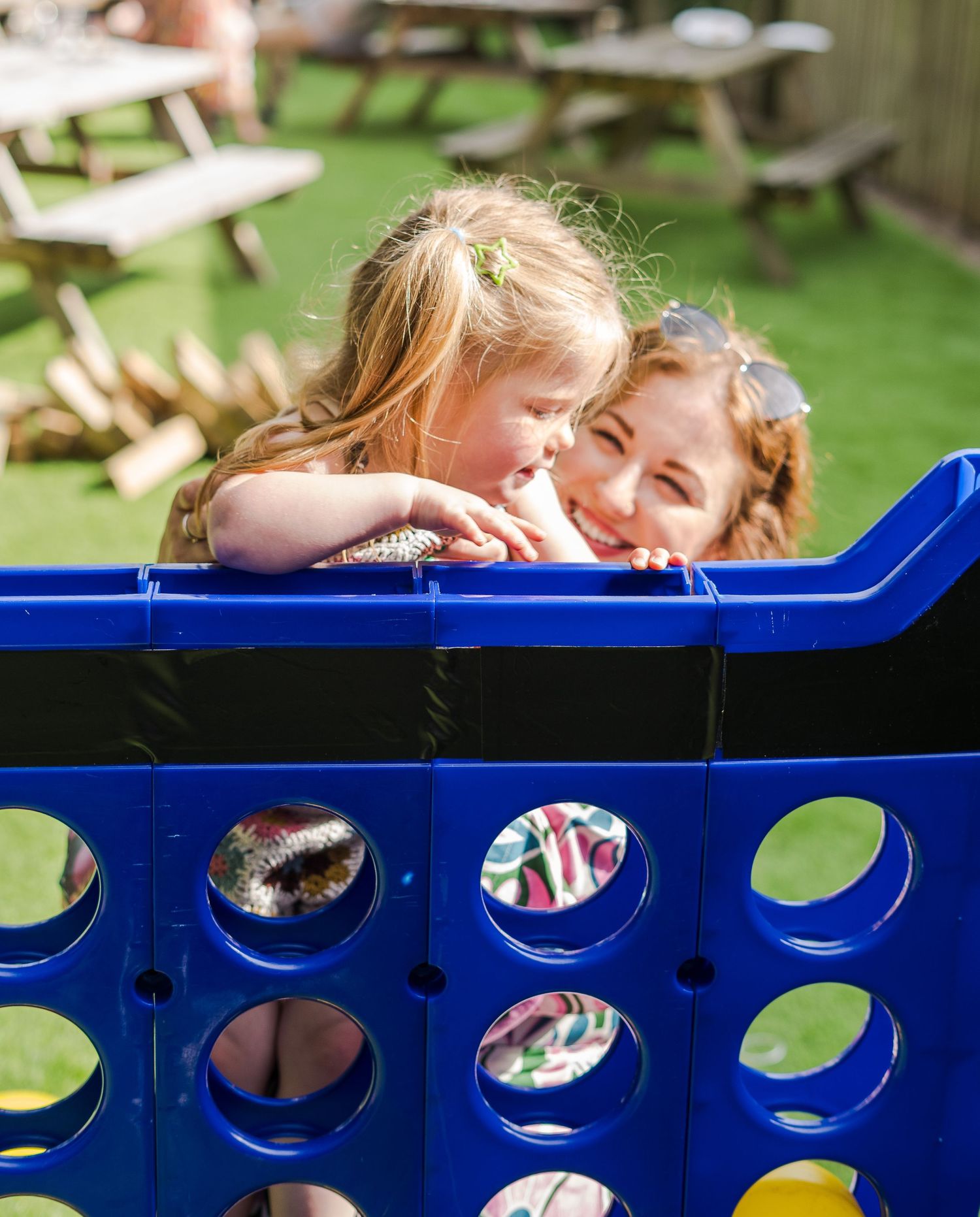 Kids playing and peeking over a blue Connect Four game board at an outdoor picnic area.