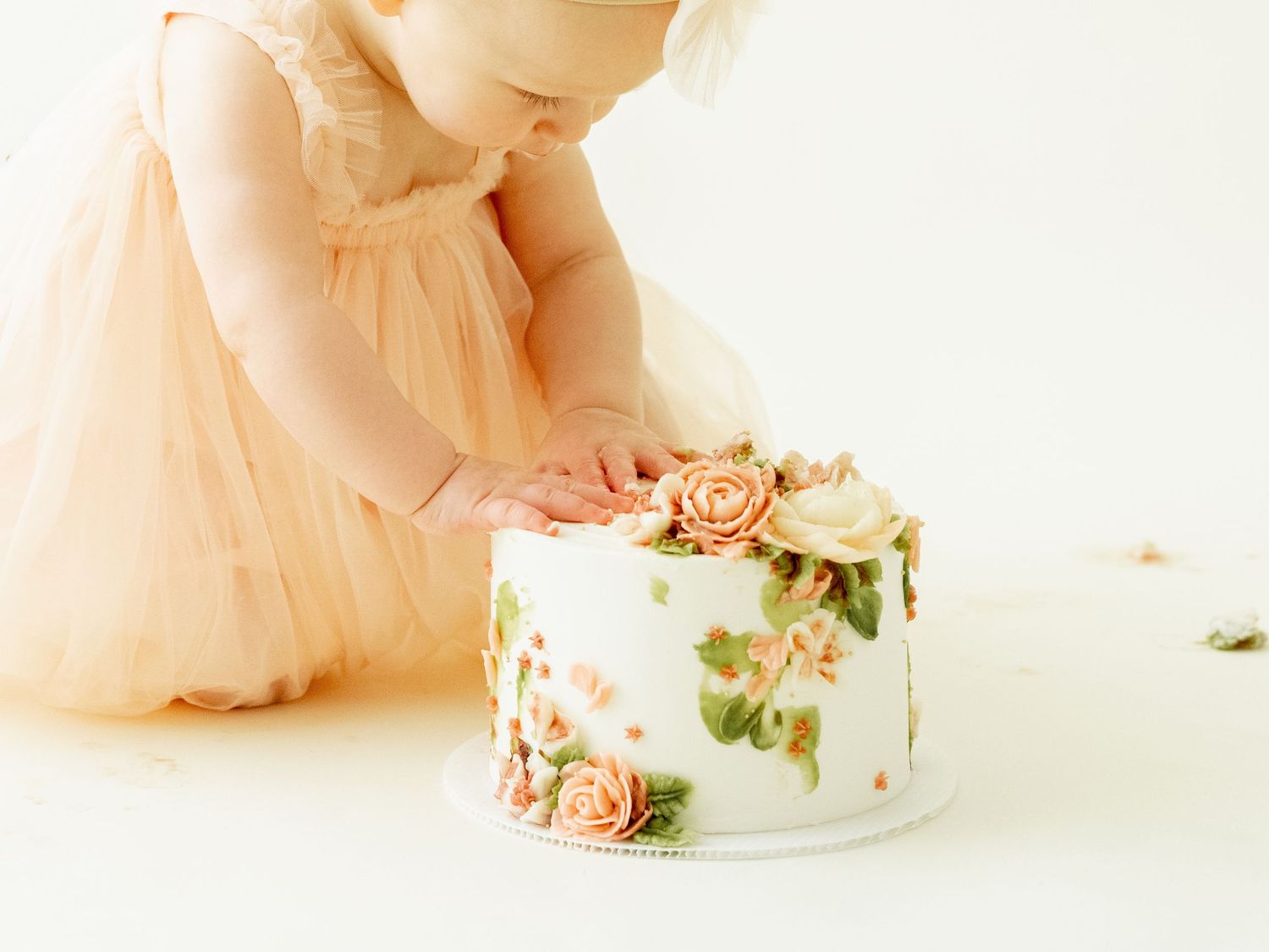 Elegant white birthday cake decorated with peach and cream roses sits next to someone wearing a tulle dress.