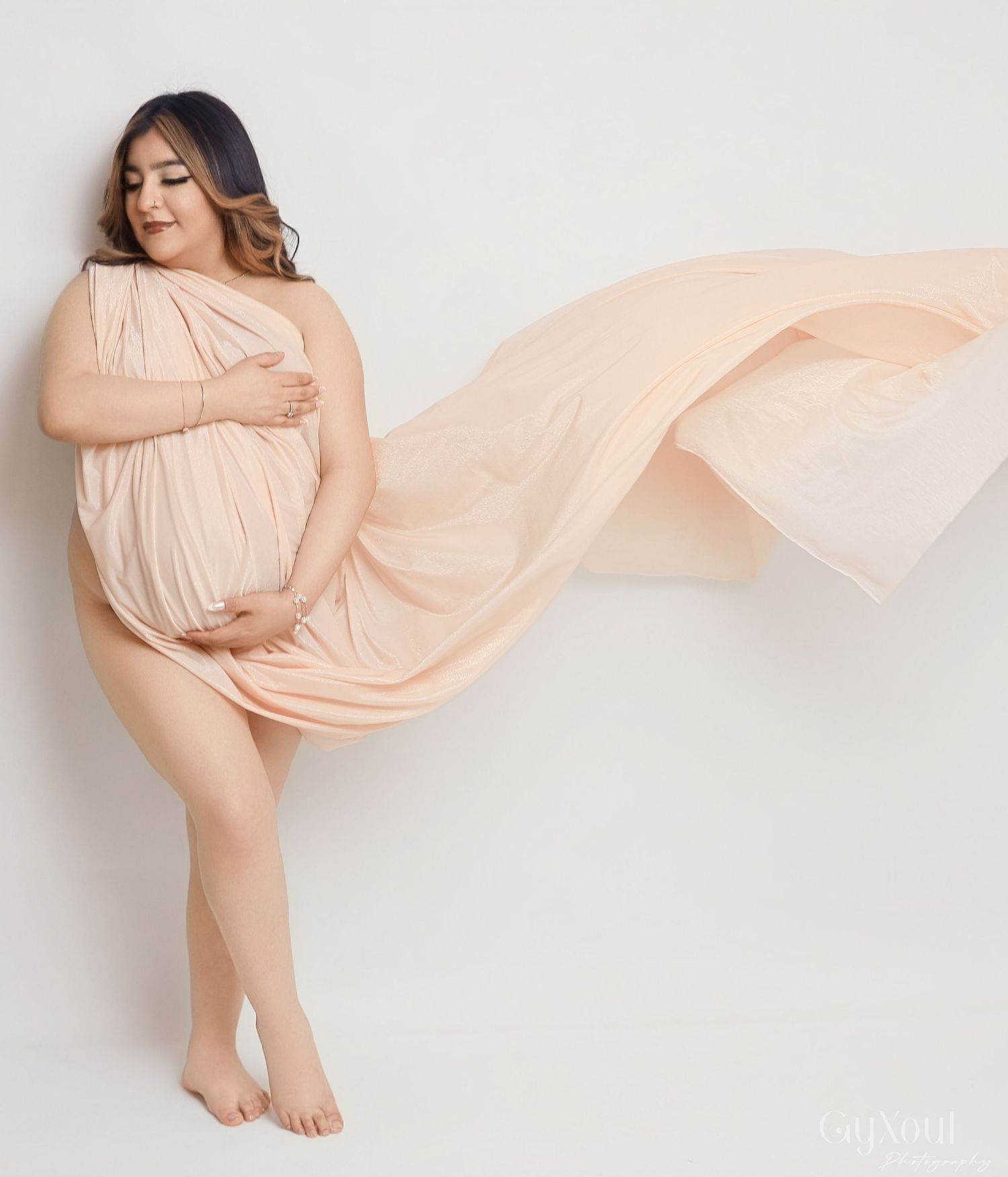 A person in a flowing peach maternity gown gracefully poses against a light backdrop during a pregnancy photoshoot.