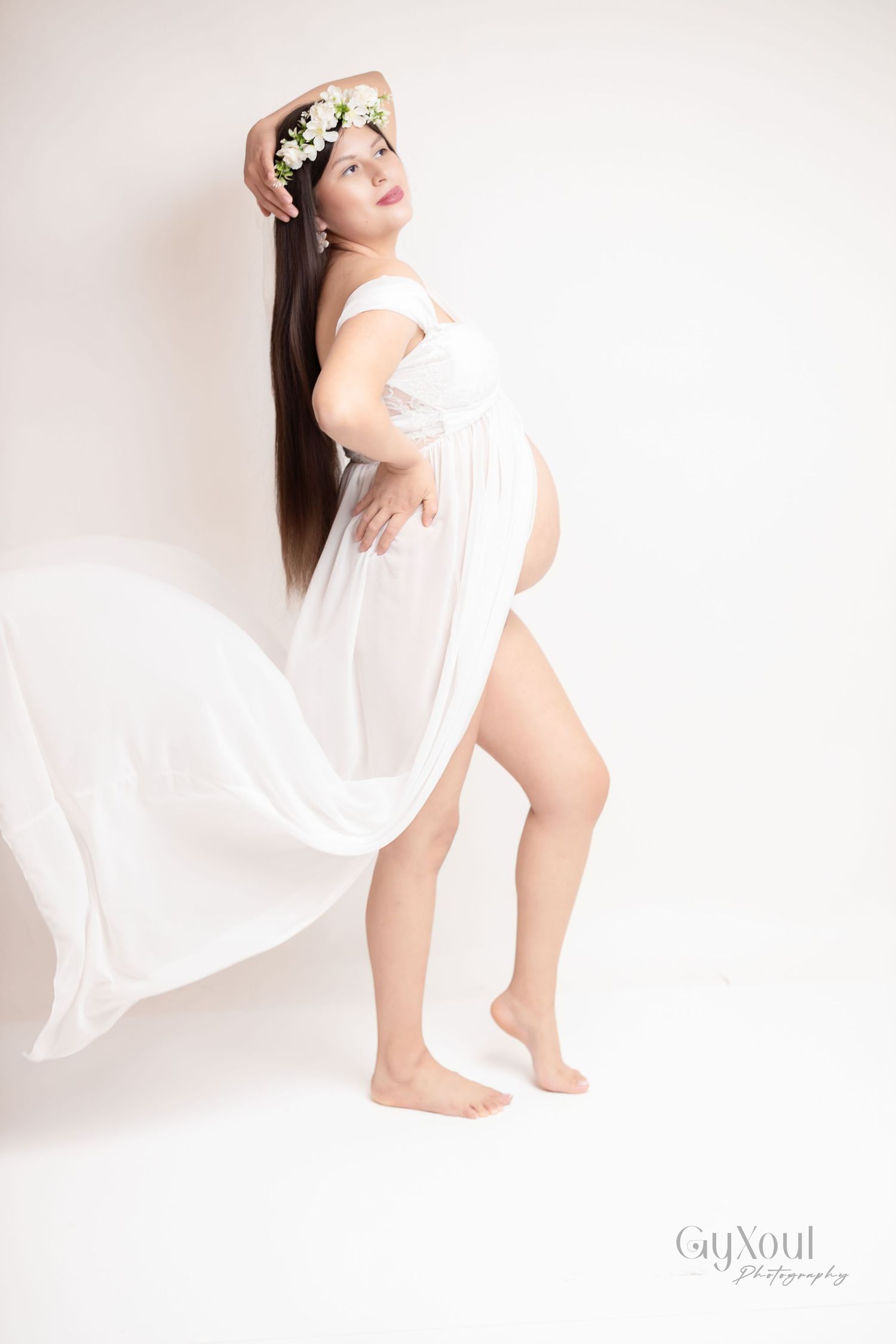 A graceful maternity portrait features a flowing white dress and floral crown against a light backdrop.