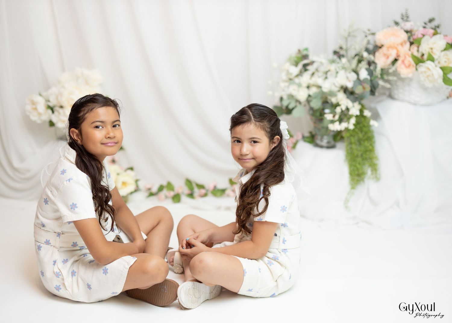 Two children in white dresses sitting on floor against white backdrop with soft floral decorations.