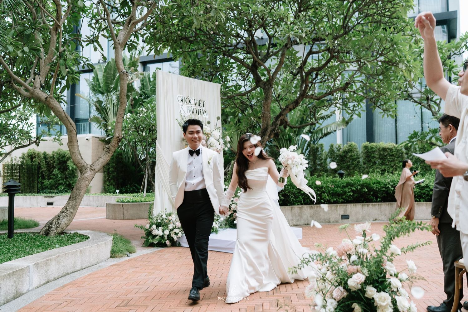 Smiling newlyweds walk hand in hand down brick aisle after outdoor garden wedding ceremony surrounded by white floral decorations.