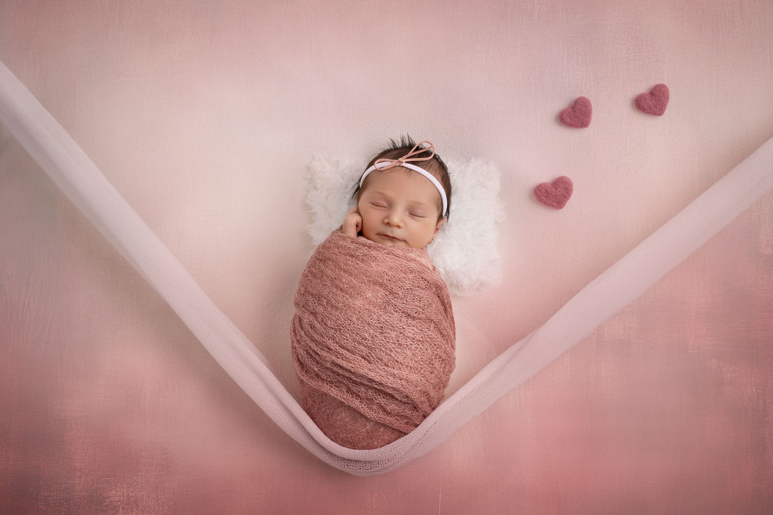 Newborn baby wrapped in pink swaddle blanket sleeps peacefully with heart decorations on pink background.