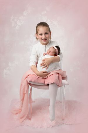 A young girl in a white top and pink skirt sits on a white chair holding a swaddled newborn against a pink background.