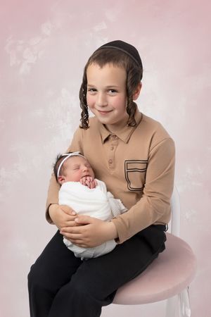 Two siblings sit together during a portrait session, with the older child holding a swaddled newborn baby on a pink chair.