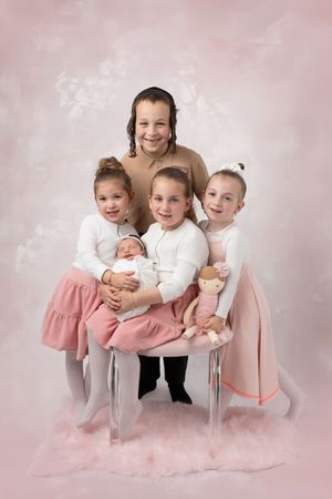 Studio portrait of children wearing white tops and pink skirts sitting together against a soft pink background.
