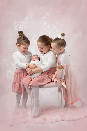 Three children in matching white tops and pink skirts sit together with a baby and doll against a pink background.