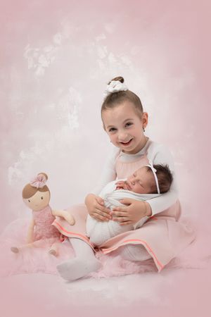 Sweet pink studio portrait with siblings in matching outfits and a plush toy doll on soft blankets.