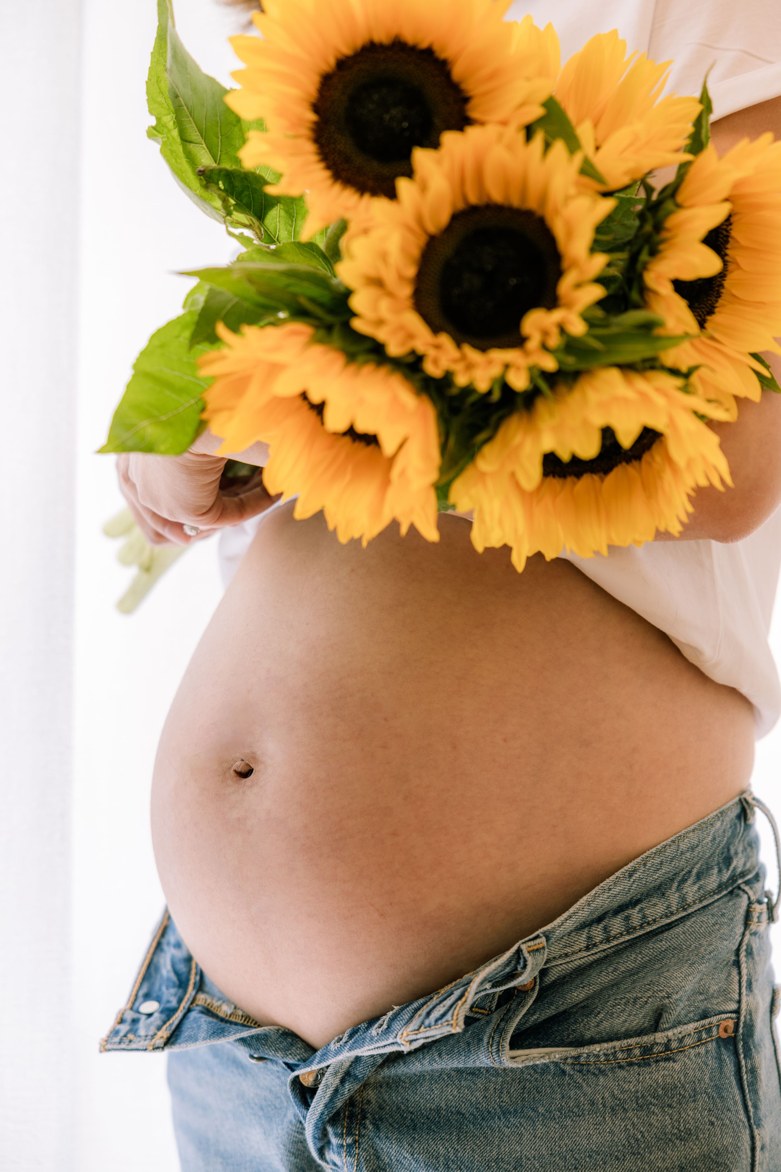 Sunflowers rest against bare pregnant belly with blue jeans.