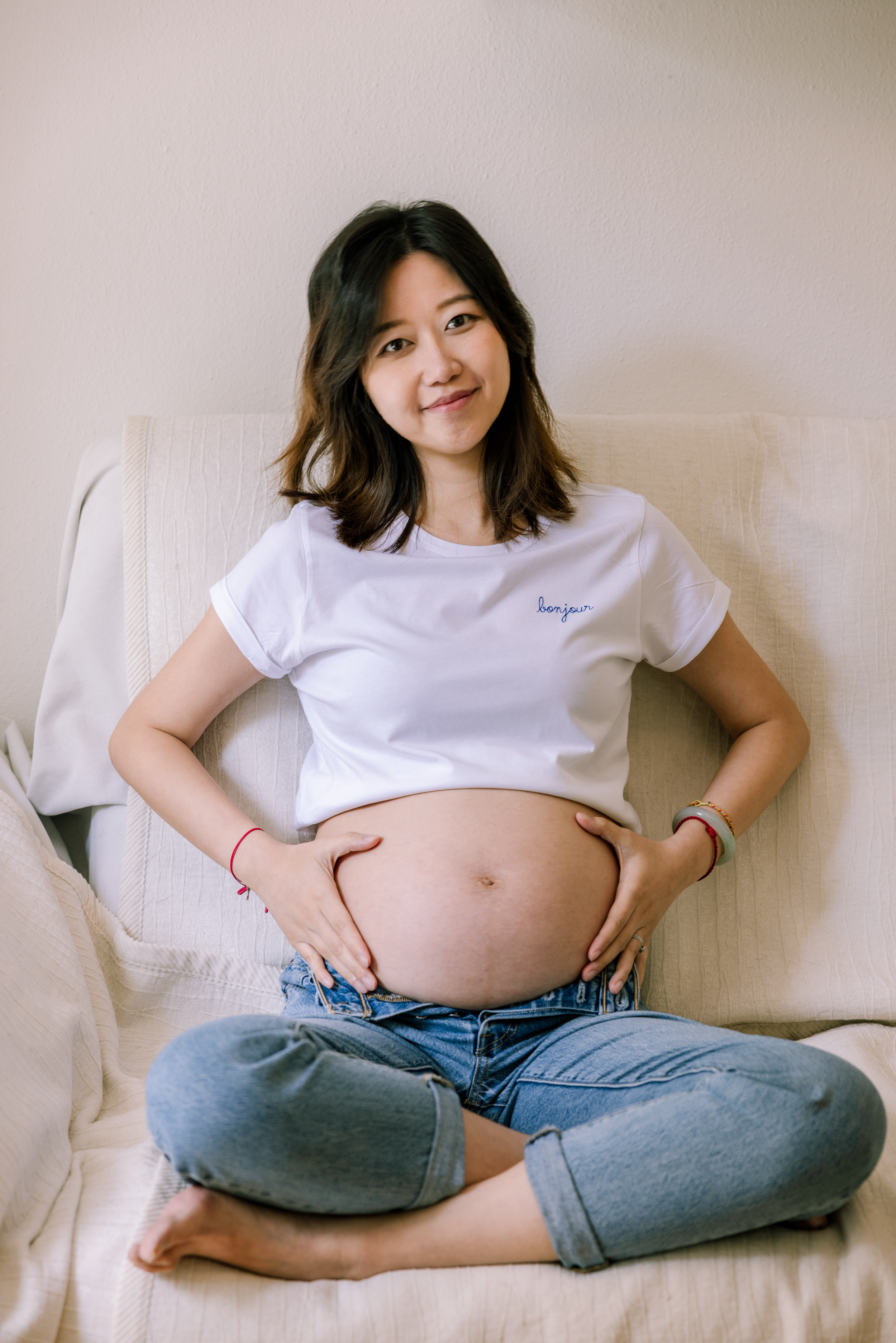 Pregnant woman in white crop top sits cross-legged on white armchair.