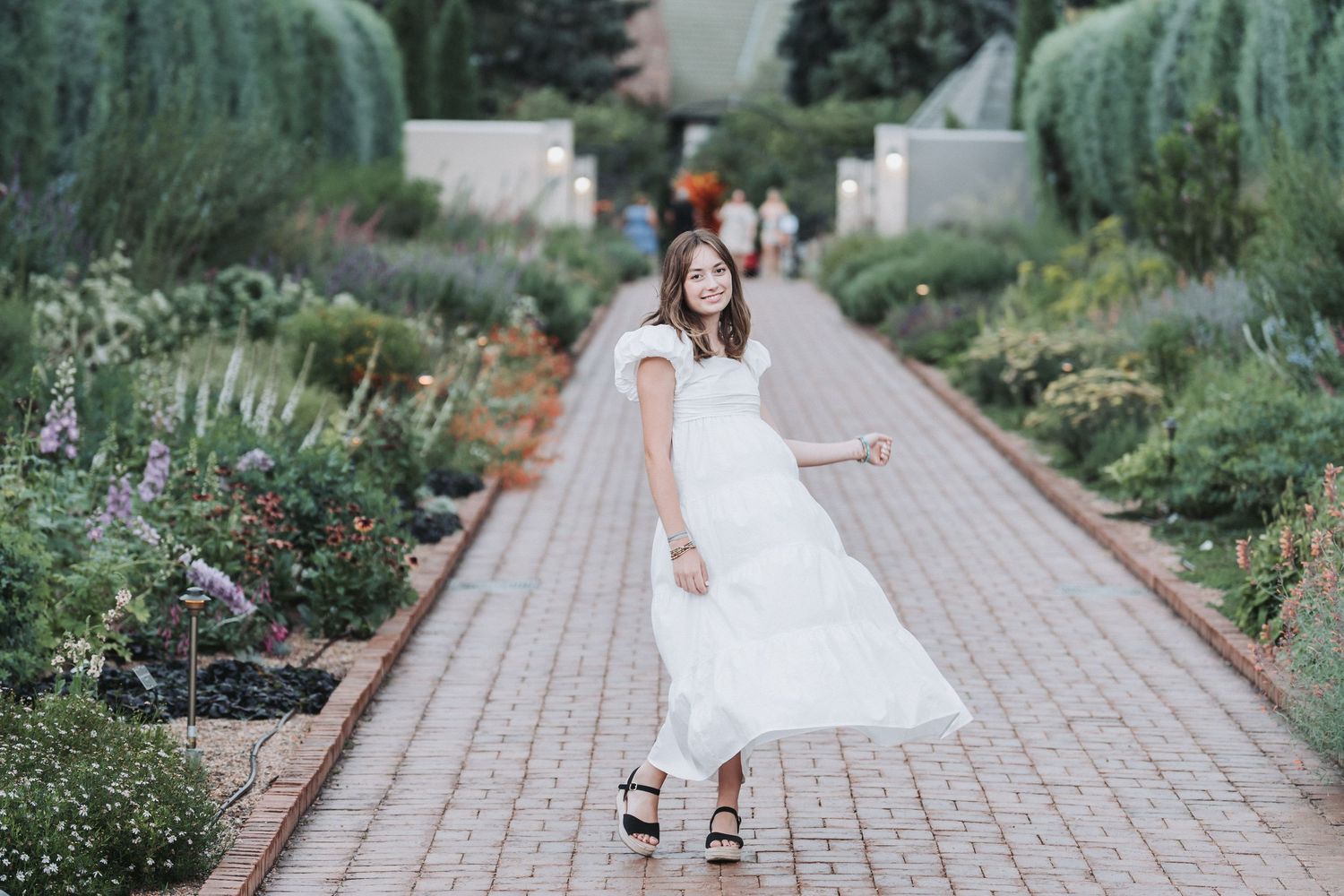 Woman in white dress twirls in formal garden pathway surrounded by greenery.