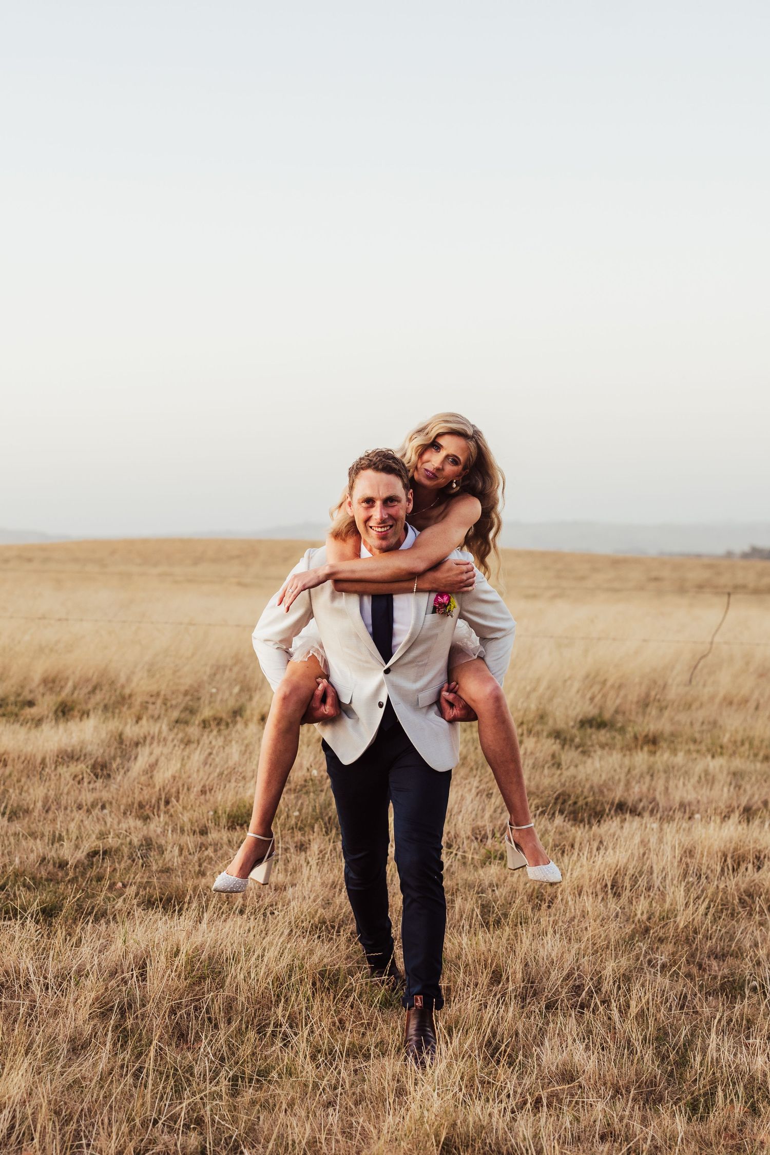 A couple shares a playful piggyback ride moment in a golden field at sunset.