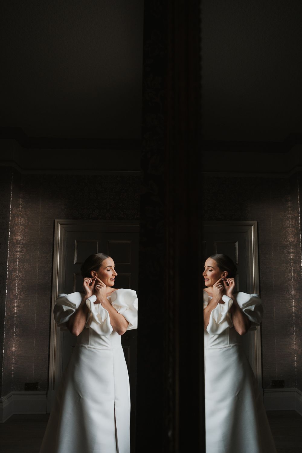 A reflection in a mirror shows an elegant figure in a white gown with dramatic sleeves in a dark moody setting.