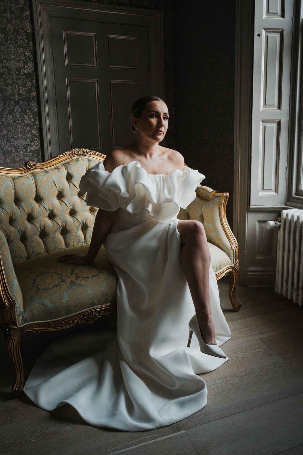 Elegant off-shoulder white wedding gown with ruffles photographed in moody lighting on vintage tufted sofa.