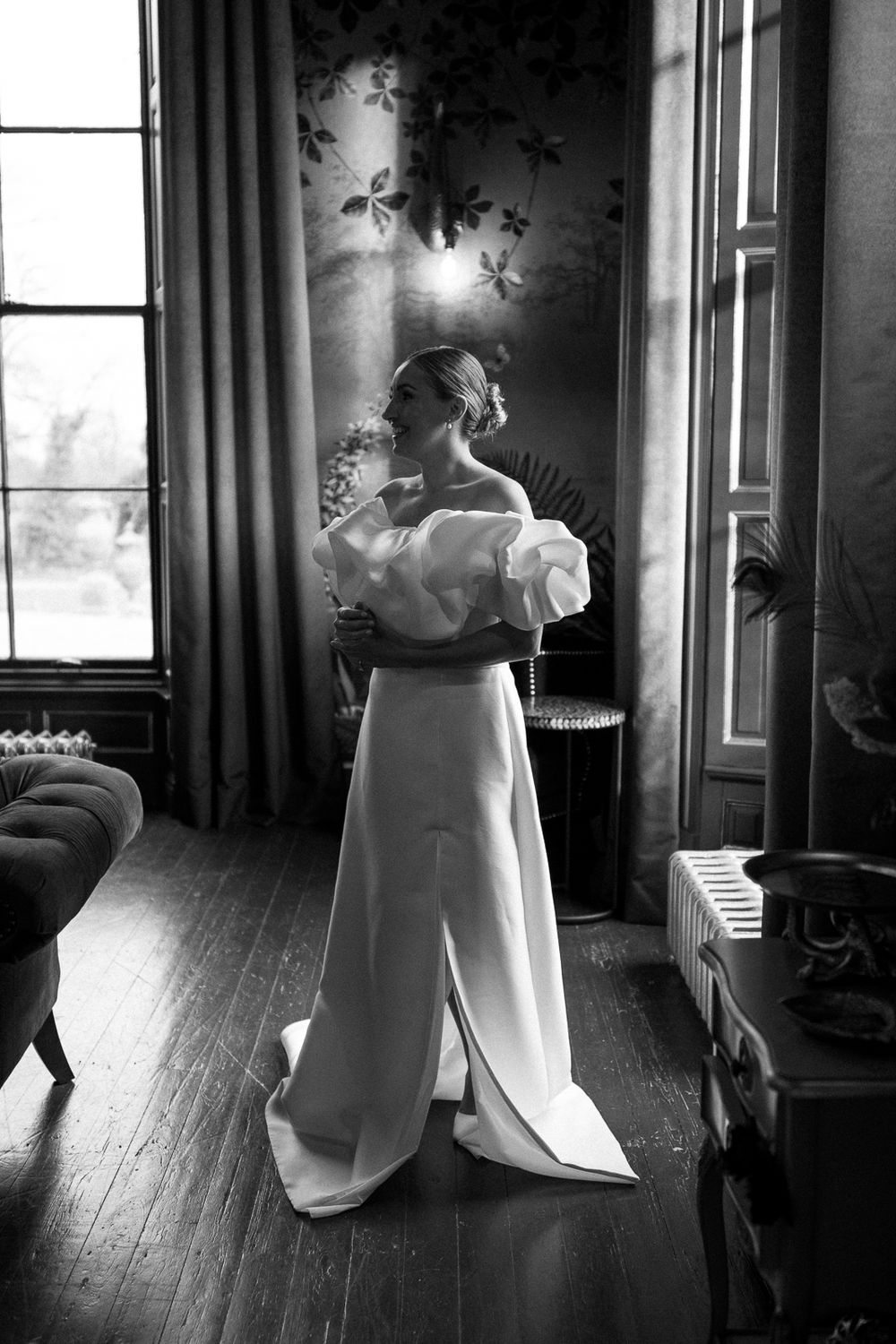 A dramatic black and white photograph of someone in an elegant white gown standing by a window with streaming sunlight.