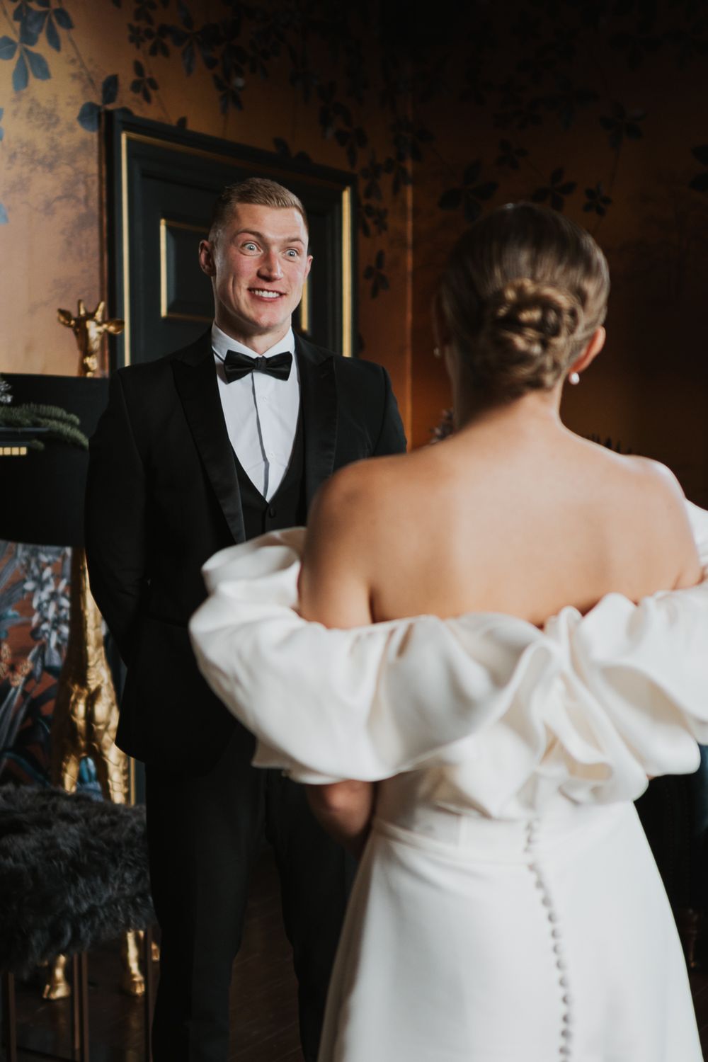 A romantic first look moment between a bride in an off-shoulder wedding dress and groom in classic black tuxedo.
