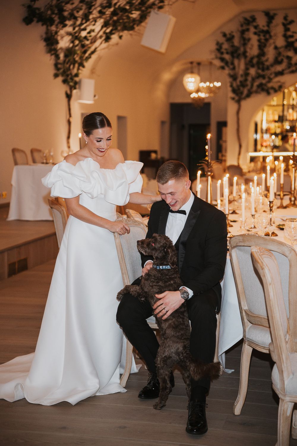 Elegant wedding couple sharing a moment with their black dog amid candlelit reception tables and arched ceilings.