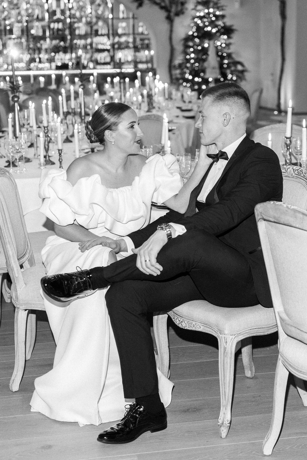 Wedding couple in formal attire sharing intimate moment at elegant candlelit table with Christmas decor in background.
