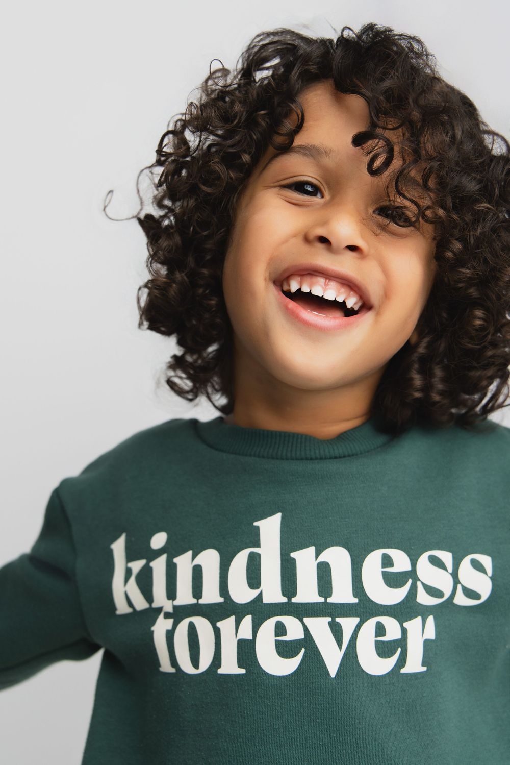 Smiling child with curly hair wearing a green sweatshirt with 'kindness forever' text print.