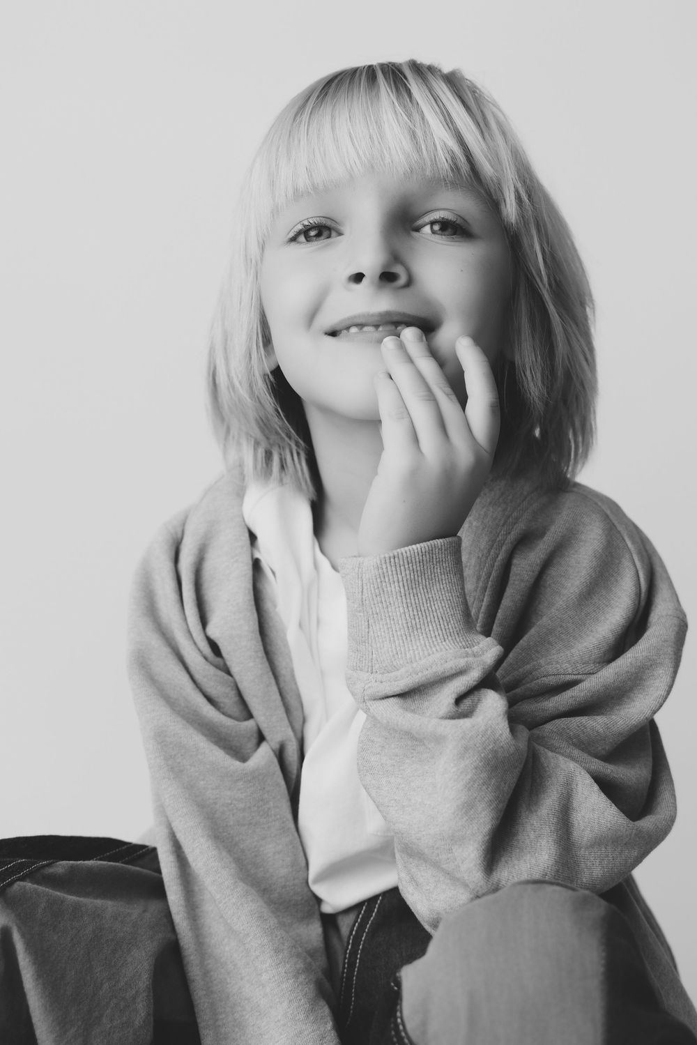 Black and white portrait of a smiling blonde child in a sweatshirt, resting chin on hand, looking up thoughtfully.