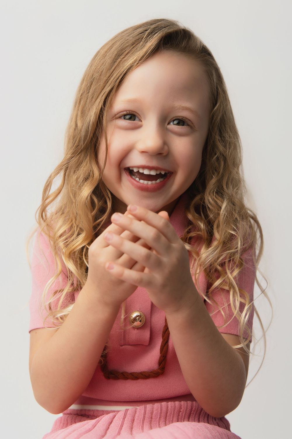 Excited young girl with curly blonde hair wearing pink, clapping hands and laughing against white background.