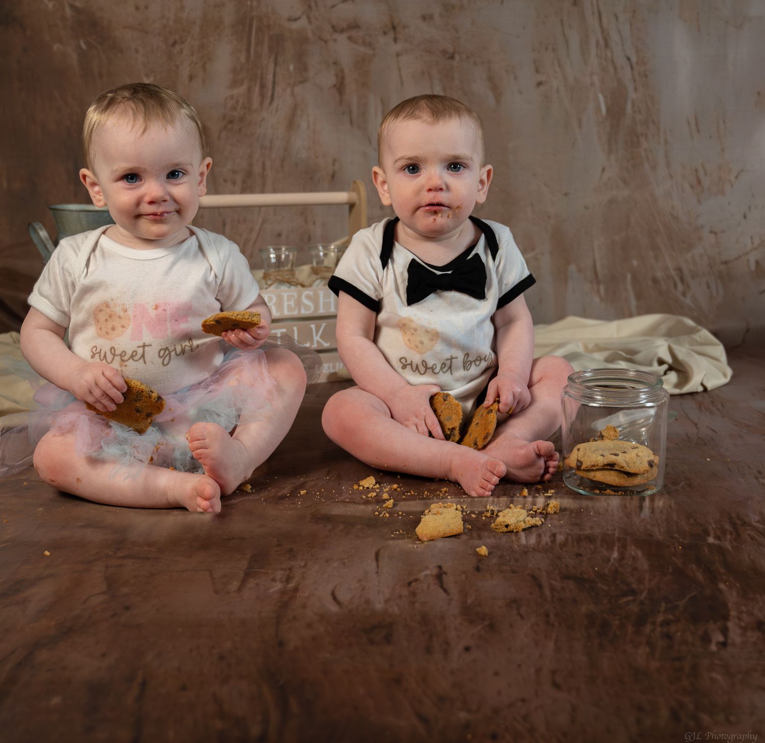 Two babies in matching outfits sit on floor surrounded by cookie crumbs during a fun first birthday smash photo session.