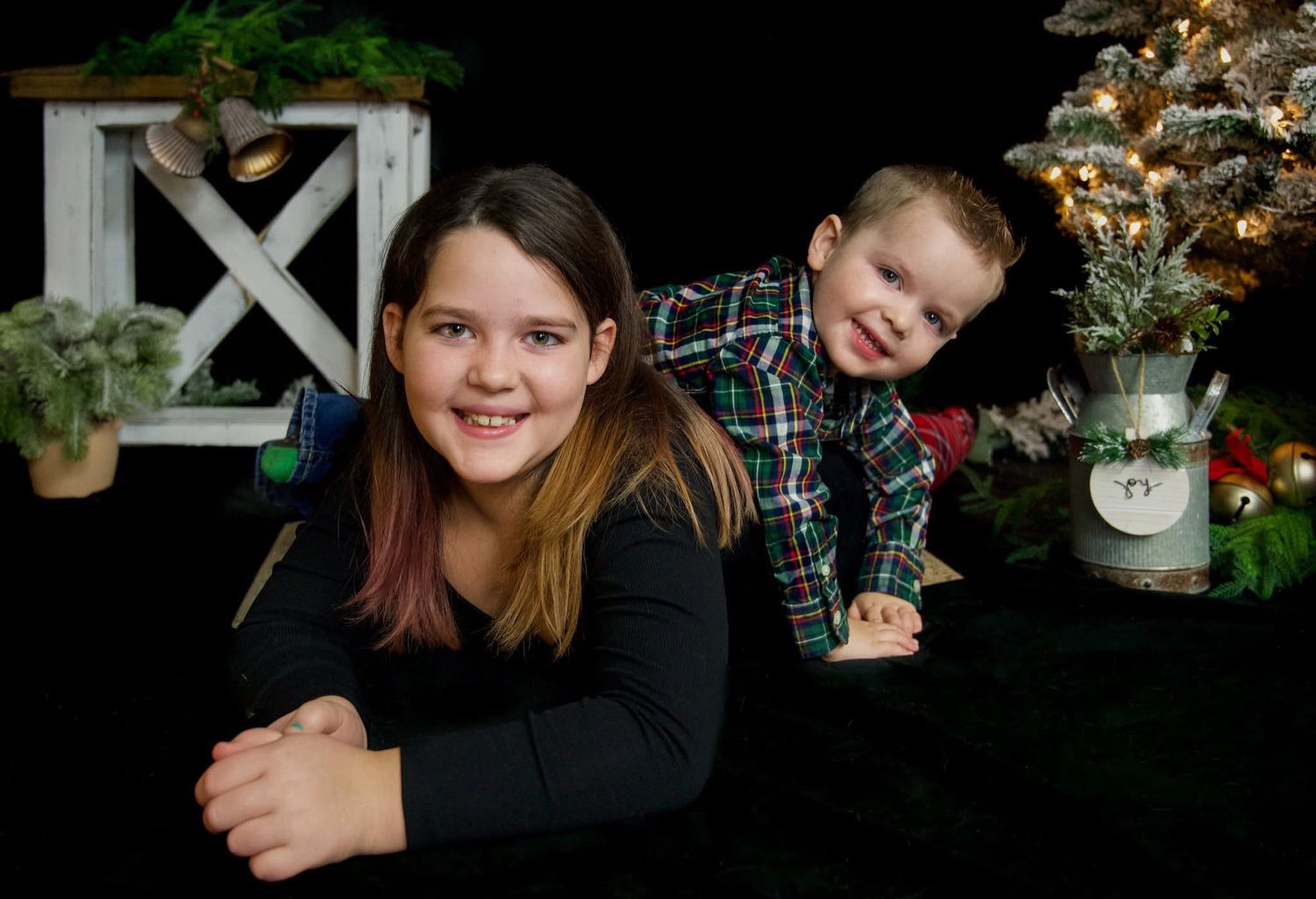 Young girl and toddler boy pose together near Christmas tree in festive holiday portrait on black background.