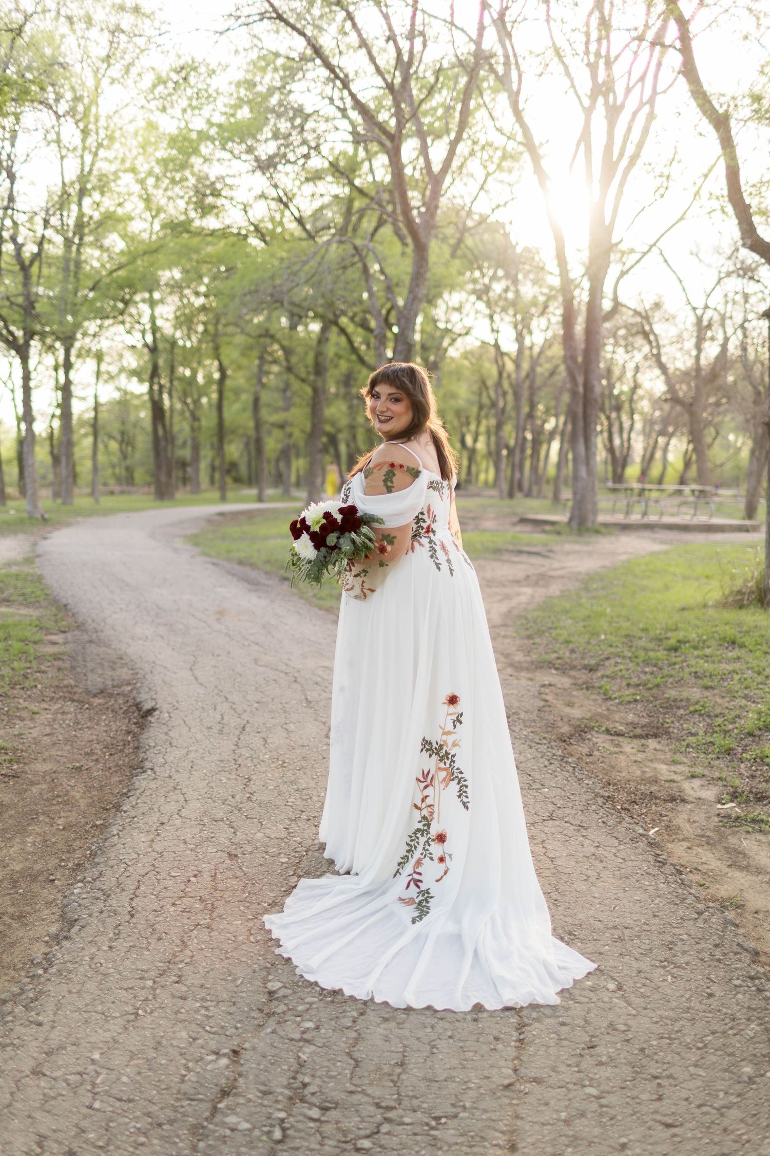 Bride in floral embroidered white gown holding bouquet on a forked path in sunlit forest at McKinney Falls State Park.