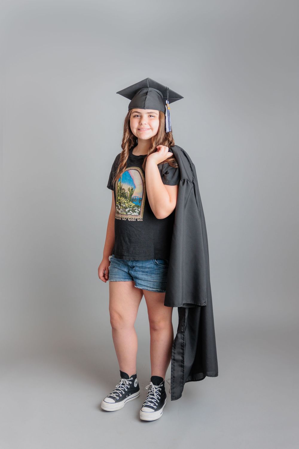 Young girl in graduation cap and gown over casual t-shirt and denim shorts, smiling for graduation photo on gray background.