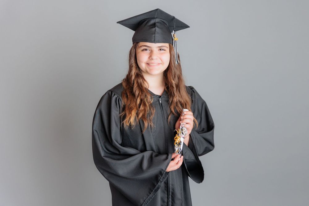 Smiling young woman in black graduation cap and gown holding diploma on gray background.