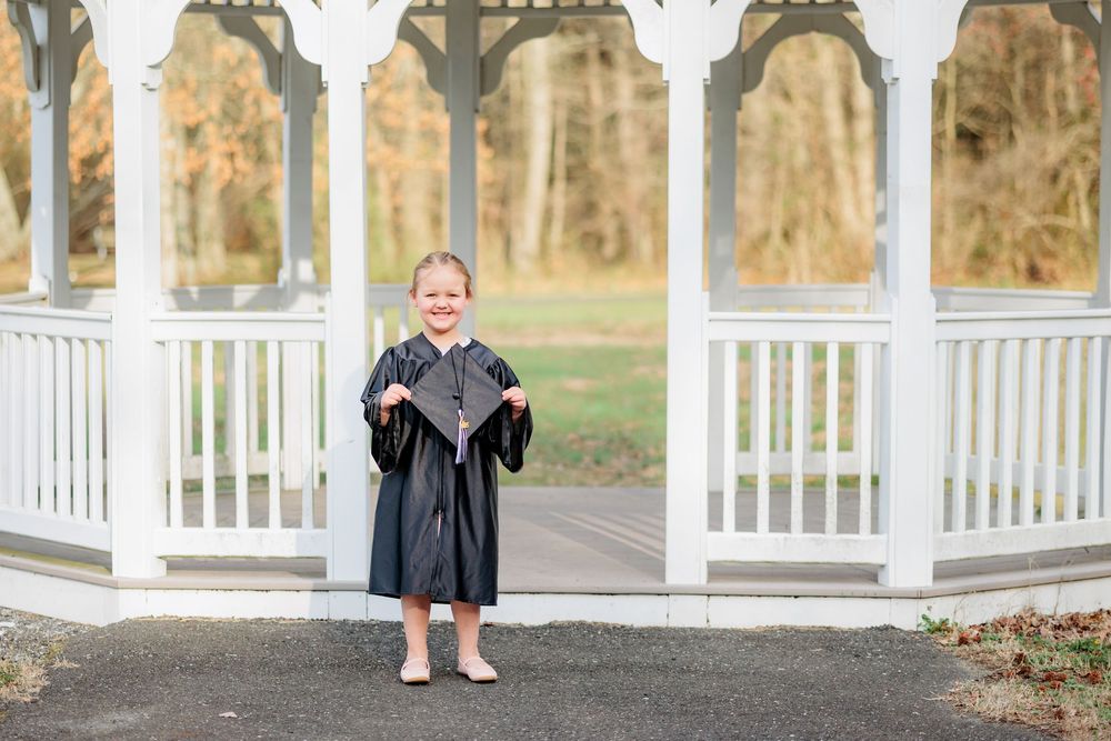 Young child in black graduation gown and cap stands proudly in front of white gazebo outdoors.