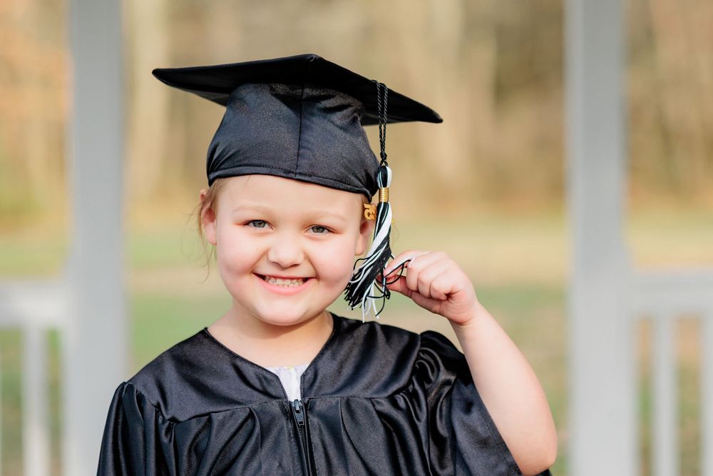 Young girl smiling in black graduation cap and gown, holding tassel, celebrating preschool or kindergarten graduation outdoors.