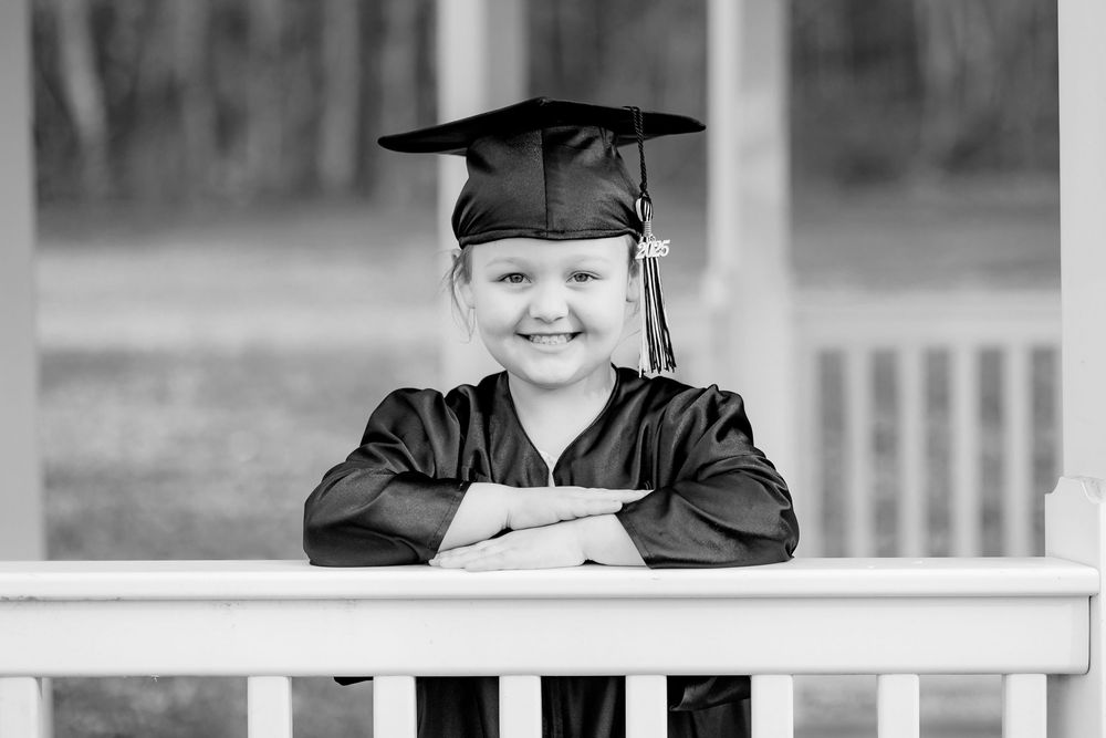 Smiling young child wearing graduation cap and gown posing on white porch in black and white photo.