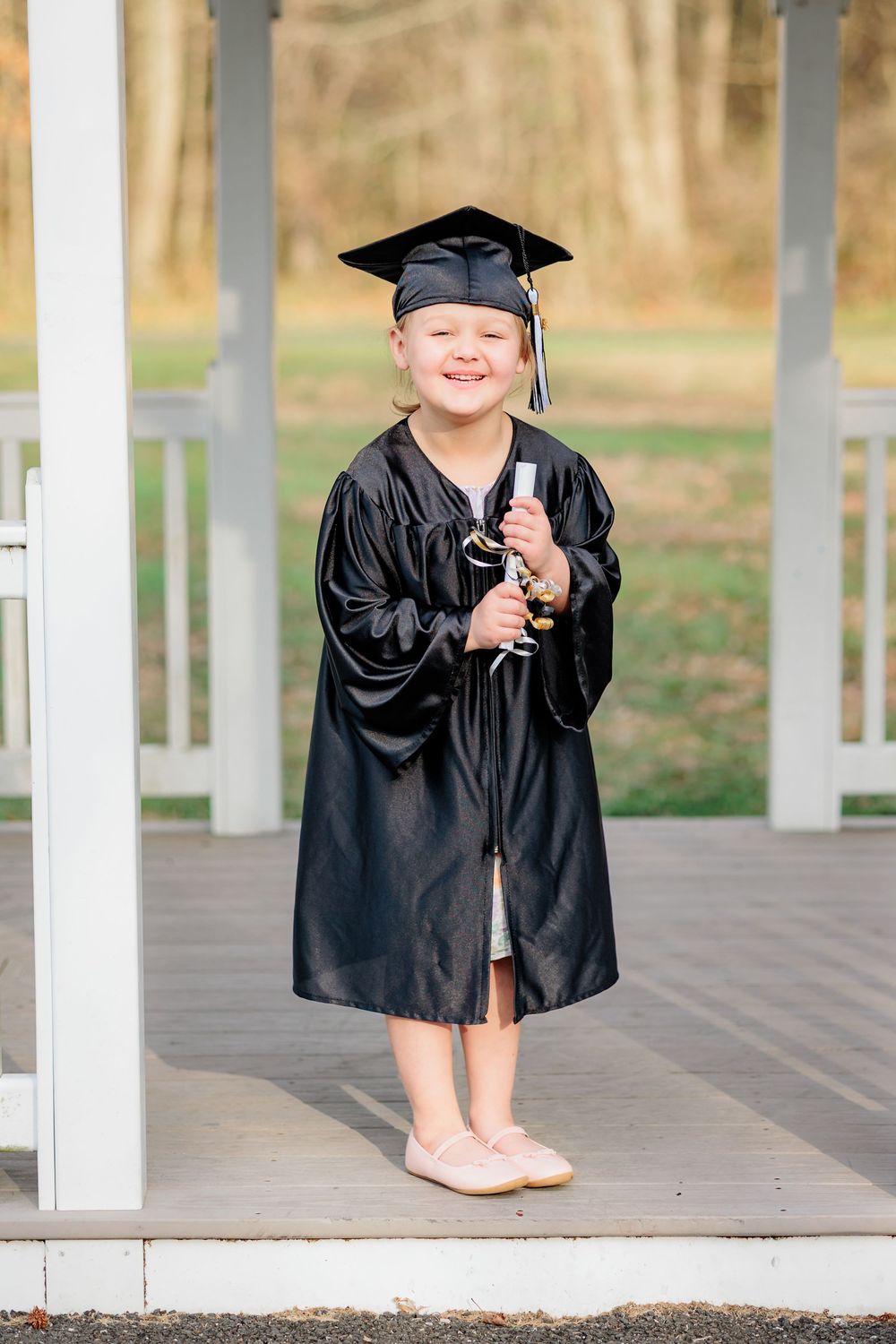 Smiling young girl in black graduation cap and gown holding diploma on porch.
