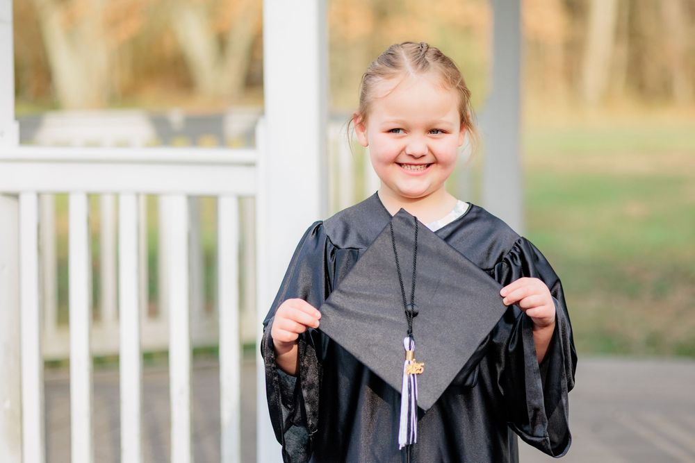 Young girl in black graduation gown and cap smiling outdoors during kindergarten graduation ceremony.