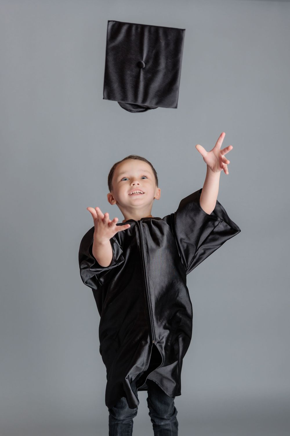 Happy young boy in black graduation gown tossing mortarboard cap in the air against gray background.