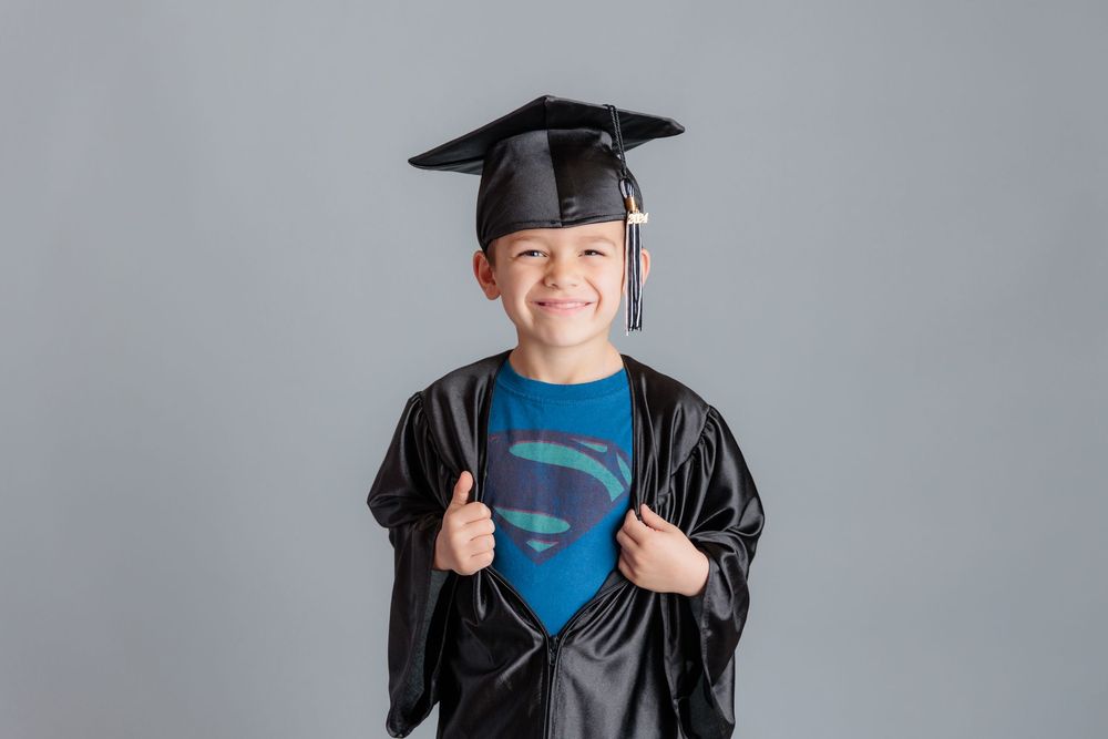 Young boy in graduation cap and gown revealing Superman shirt, smiling and giving thumbs up against gray background.