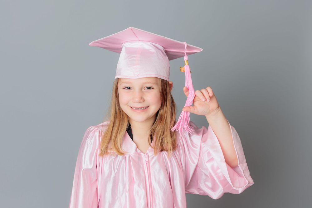 Young girl smiling in pink graduation cap and gown, holding tassel, on gray background.
