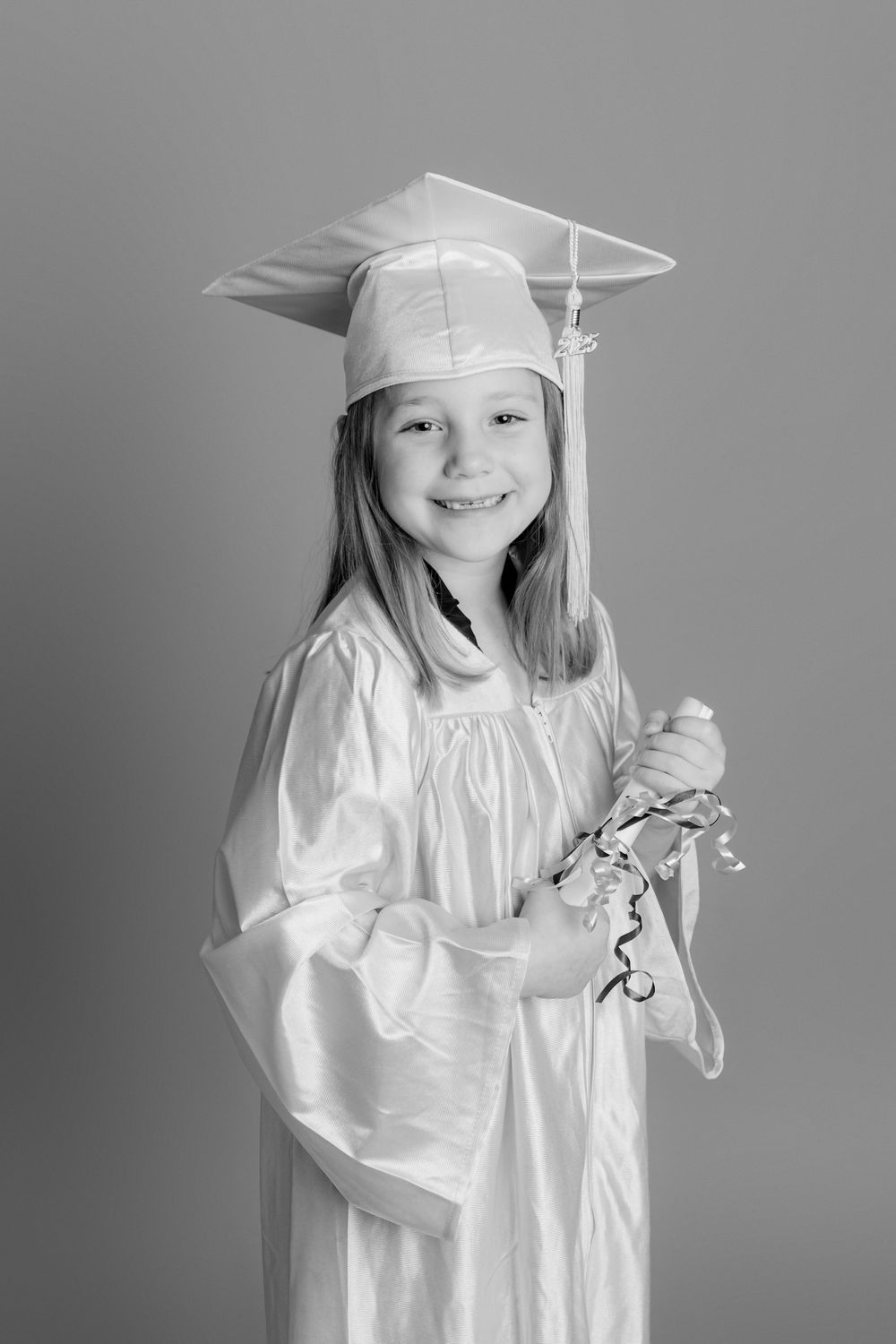 Young girl smiling in white graduation cap and gown holding ribbons, black and white photo.