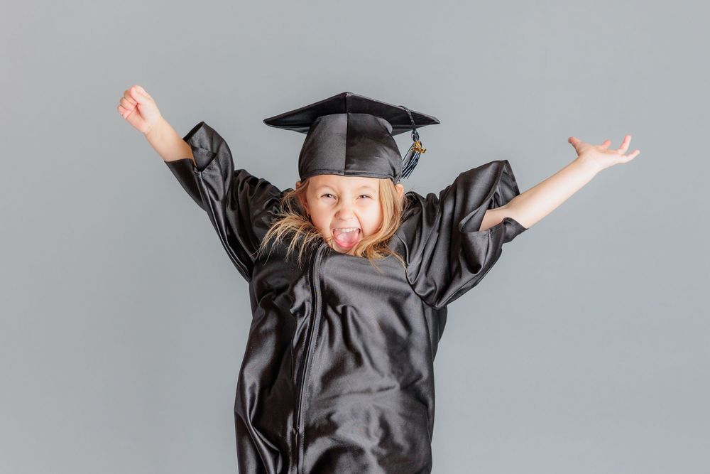 Excited young girl in black graduation cap and gown celebrating with arms raised and tongue out.