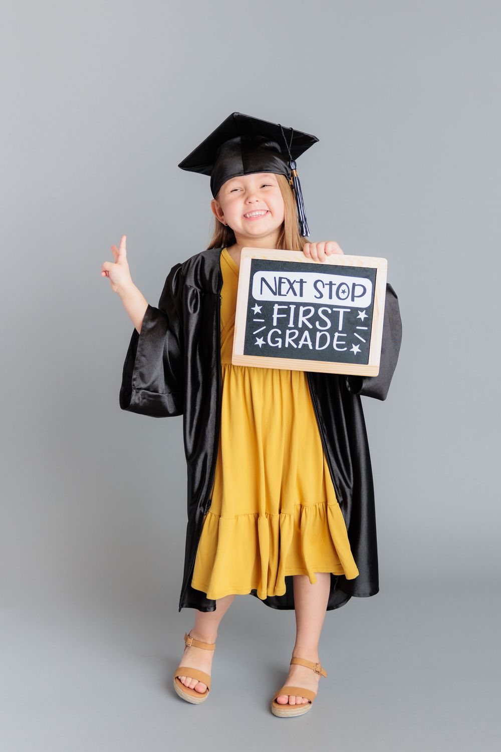 Smiling girl in graduation cap and gown holds a chalkboard sign reading Next Stop First Grade.
