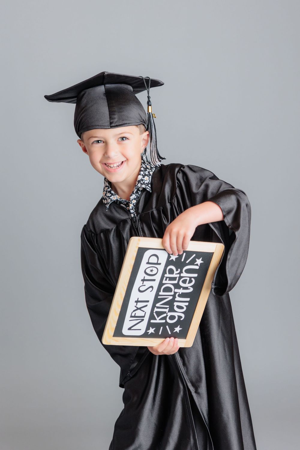 Young child in black graduation cap and gown holding a chalkboard sign reading Next Stop Kindergarten.