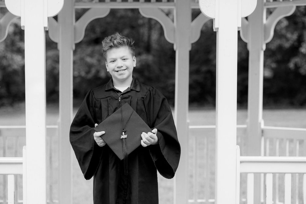 Smiling graduate in black gown holding cap outdoors near white gazebo in black and white photo.