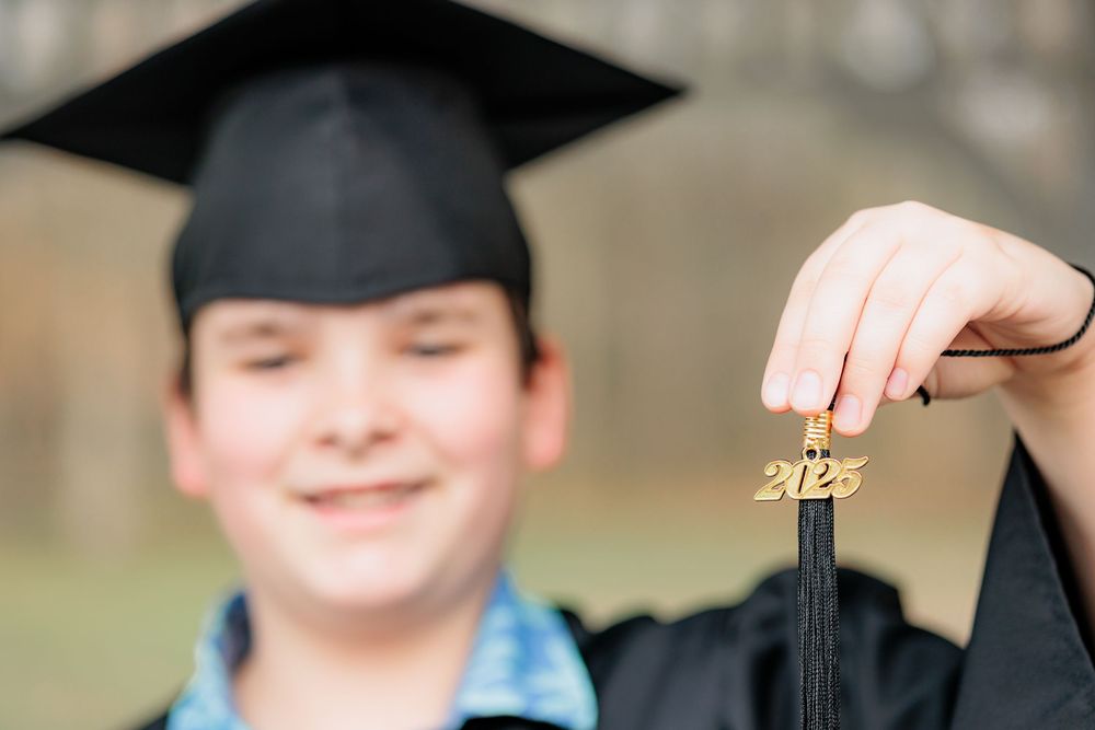 Young graduate in black cap and gown smiling while holding a gold 2023 tassel charm at graduation.
